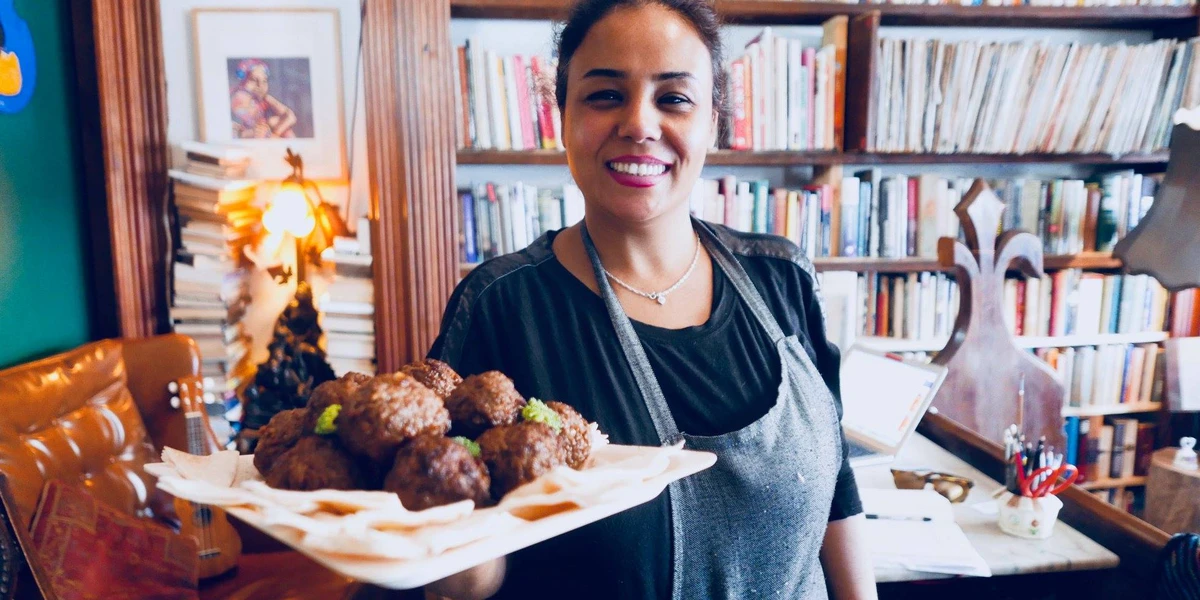 A woman wearing an apron smiles while holding a plate of meatballs covered with a white napkin. Shelves filled with books and a decorative lamp are visible in the background.