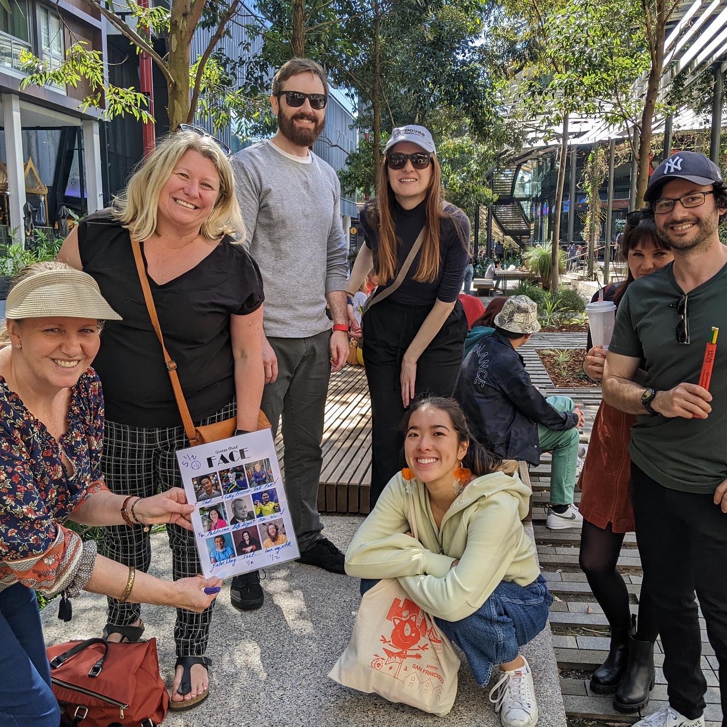 A group of seven people, six standing and one kneeling, smile at the camera in an outdoor setting with trees and modern buildings. One individual holds up a collage. They appear relaxed and casually dressed, enjoying a sunny day together. A group of seven people, six standing and one kneeling, smile at the camera in an outdoor setting with trees and modern buildings. One individual holds up a collage. They appear relaxed and casually dressed, enjoying a sunny day together.