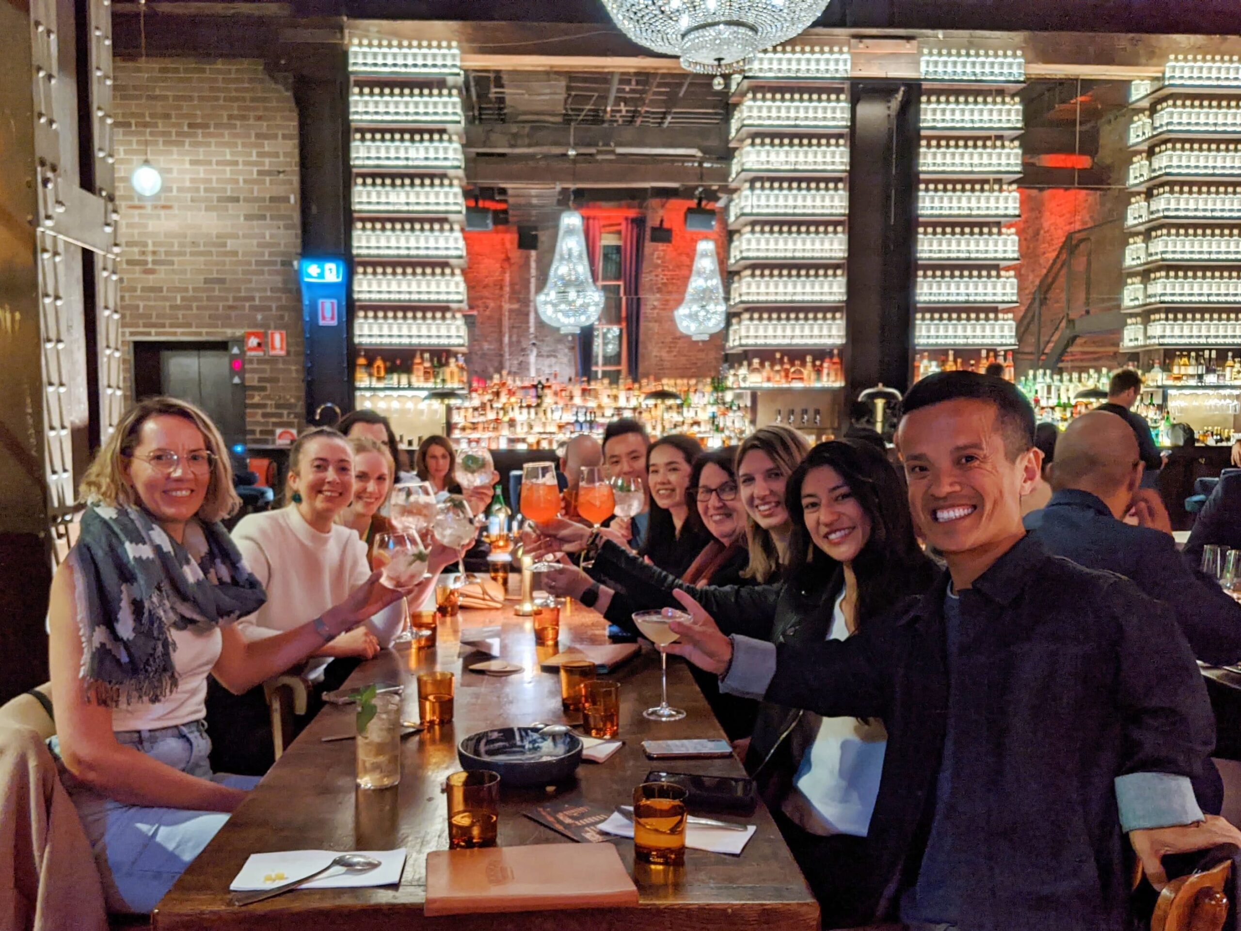 A group of people smiling and raising glasses during a gathering at a dimly lit bar with vivid lights illuminating a large chandelier, shelves of bottles, and brick walls in the background. They are seated around a wooden table with drinks and menus. A group of people smiling and raising glasses during a gathering at a dimly lit bar with vivid lights illuminating a large chandelier, shelves of bottles, and brick walls in the background. They are seated around a wooden table with drinks and menus.