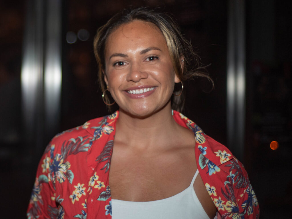 Person smiling at the camera, wearing a red floral-patterned shirt over a white top. The background is dimly lit, emphasizing the person's smile. They have hoop earrings and their hair is pulled back.