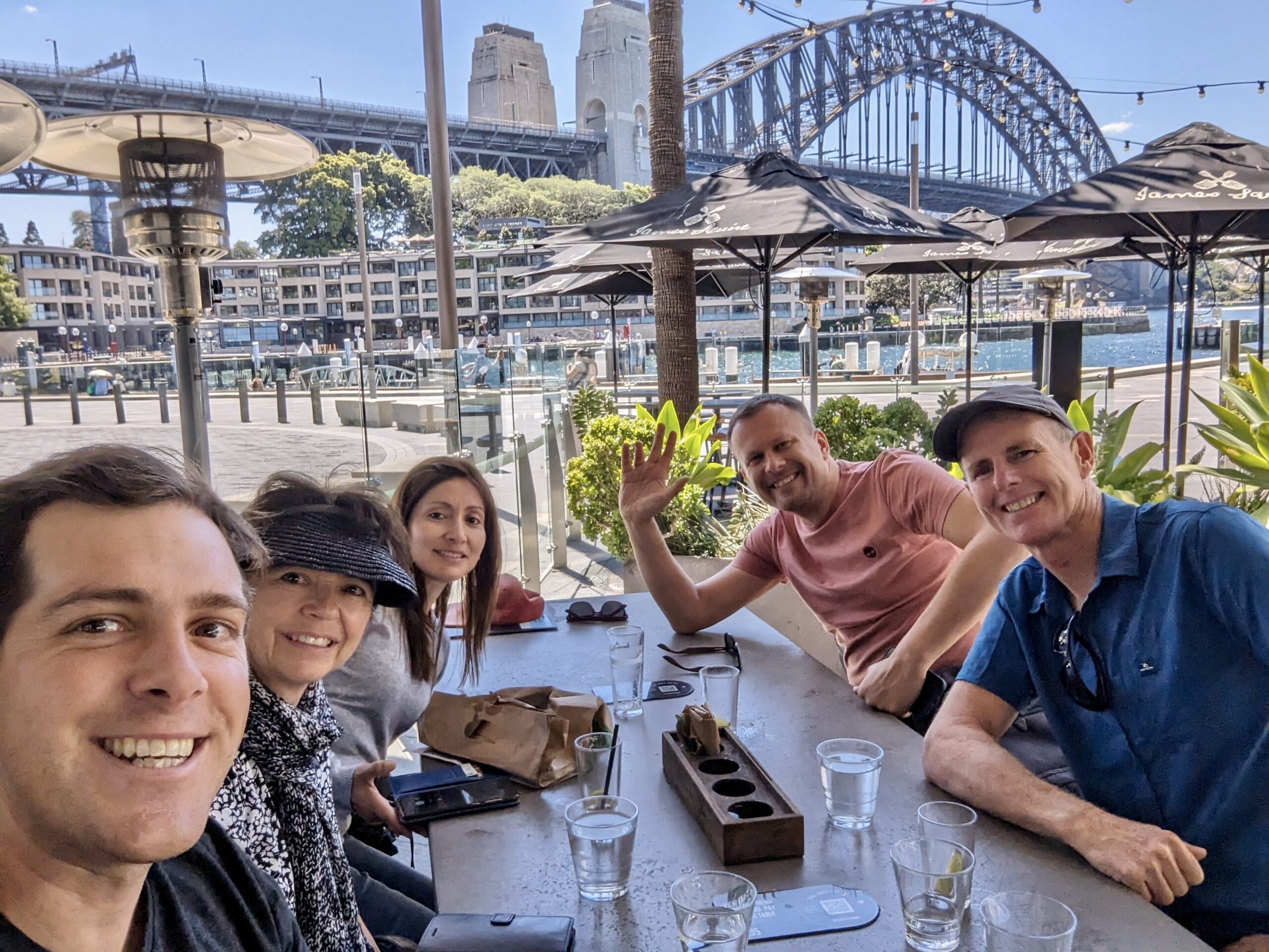 Five people sit at an outdoor dining table, smiling towards the camera with the Sydney Harbour Bridge in the background. Drinks and a small bag are on the table. It's a sunny day, and they are seated under umbrellas. Five people sit at an outdoor dining table, smiling towards the camera with the Sydney Harbour Bridge in the background. Drinks and a small bag are on the table. It's a sunny day, and they are seated under umbrellas.