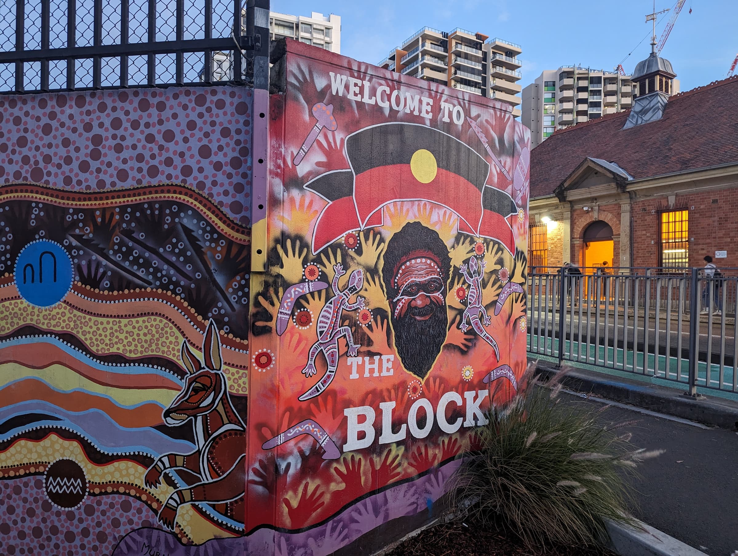 A vibrant mural welcomes visitors to "The Block" with Aboriginal art, including traditional patterns, animals, and dancing figures. The mural prominently features a central figure with the Aboriginal flag overhead. Nearby, urban buildings and an older brick structure set the perfect backdrop for a bar crawl. A vibrant mural welcomes visitors to "The Block" with Aboriginal art, including traditional patterns, animals, and dancing figures. The mural prominently features a central figure with the Aboriginal flag overhead. Nearby, urban buildings and an older brick structure set the perfect backdrop for a bar crawl.