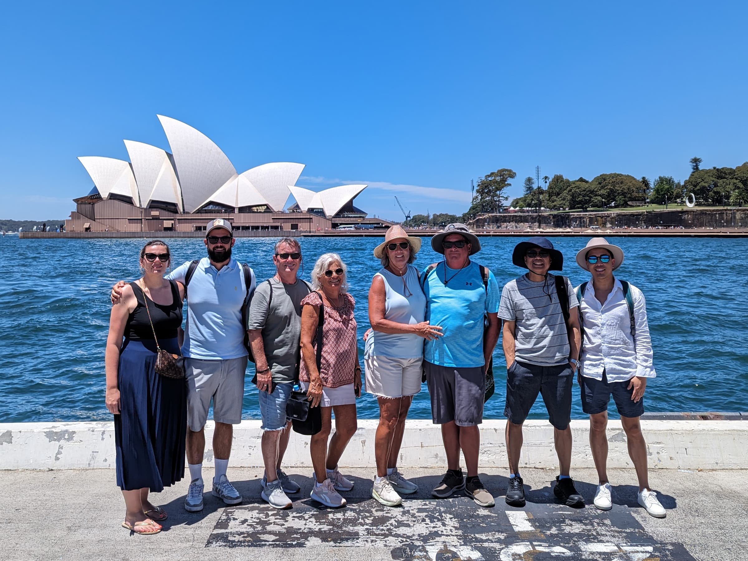 A group of eight people stands together along the waterfront with the Sydney Opera House in the background. The group, casually dressed and wearing hats and sunglasses under a clear, sunny sky, appears to be enjoying one of those delightful Local Sauce Tours.